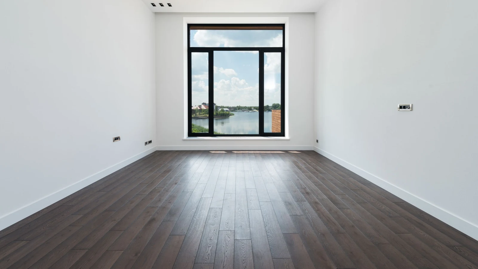 Pièce vide aux murs blancs et au parquet foncé, grande fenêtre au fond